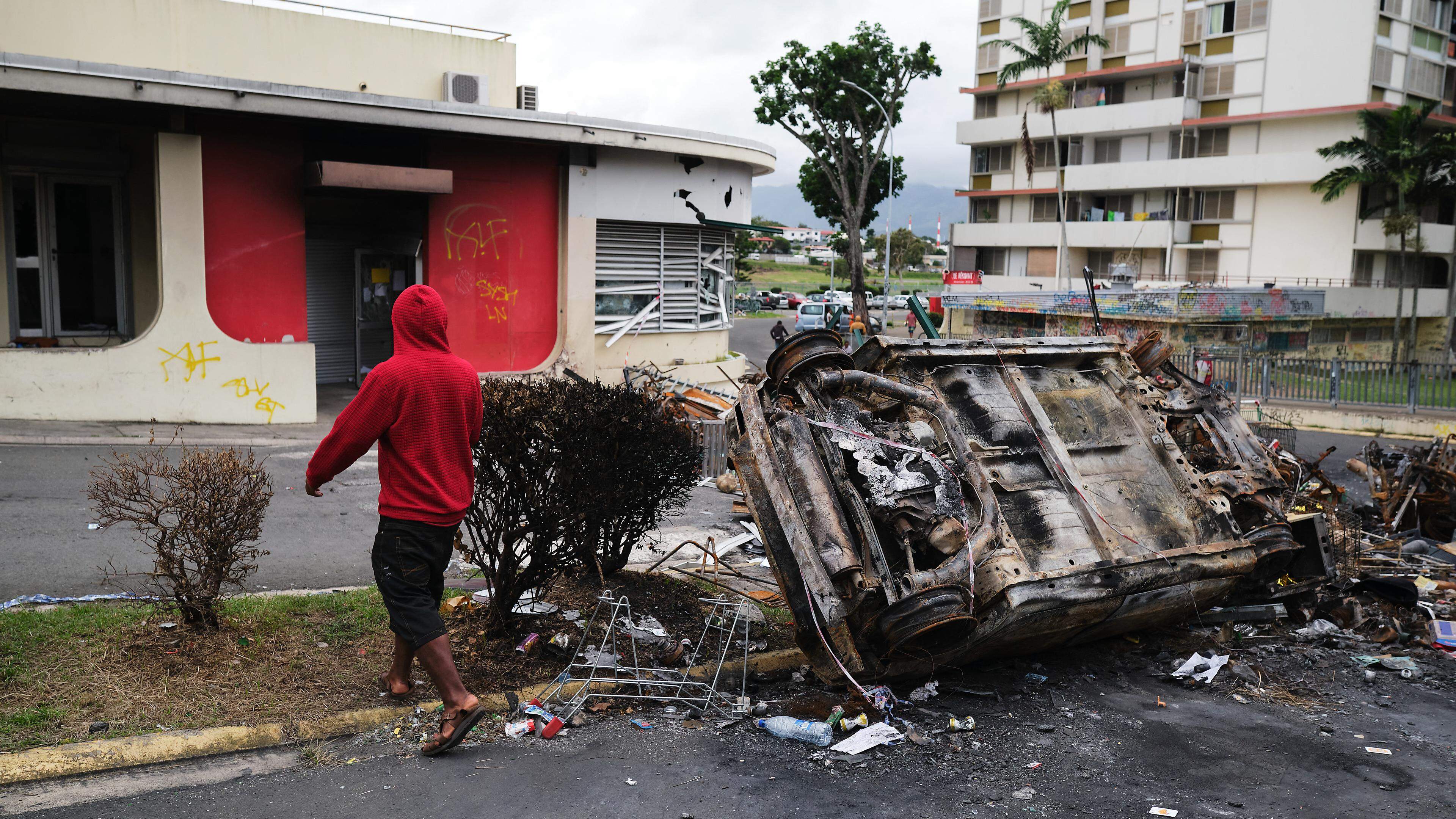 A man walks past a burnt vehicle at an independantist roadblock at Magenta Tour district in Noumea, France's Pacific territory of New Caledonia, on May 22, 2024. The Pacific territory of 270,000 people has been in turmoil since May 13, when violence erupted over French plans to impose new voting rules that would give tens of thousands of non-indigenous residents voting rights. The unrest has left six people dead, including two police, and hundreds injured. (Photo by Theo Rouby / AFP)